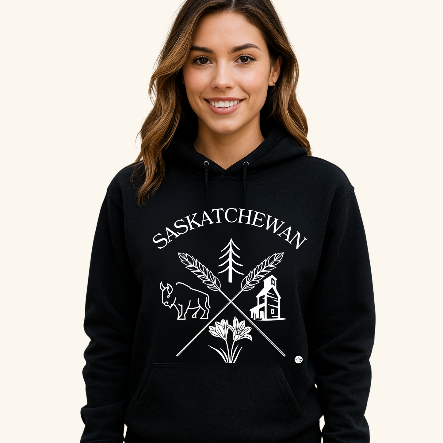 A young man with short dark hair wears a black hoodie featuring a white “Saskatchewan Prairie Roots” design with symbols of prairie life, including a bison, tree, wheat, and grain elevator. He stands with a neutral expression against a soft cream-colored background.