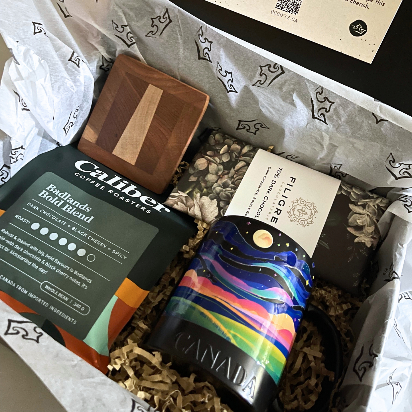 Full view of the Sunrise on the Prairie Box, showing all items inside a black magnetic gift box with a welcome card attached to the lid. Contents include Caliber Coffee, Filigree chocolate bar, a handmade wooden coaster, and a Northern Lights mug. Tissue paper with prairie motifs lines the box.