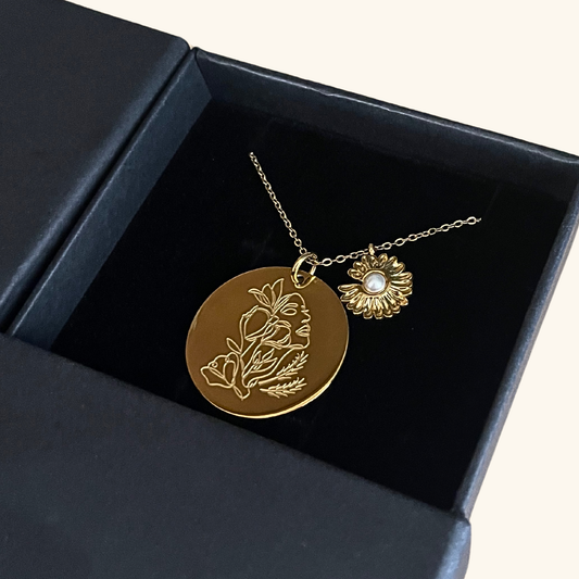 A gold necklace displayed in a black gift box, featuring a round pendant engraved with the silhouette of a woman intertwined with prairie flowers, alongside a sunflower charm with a pearl-like center.