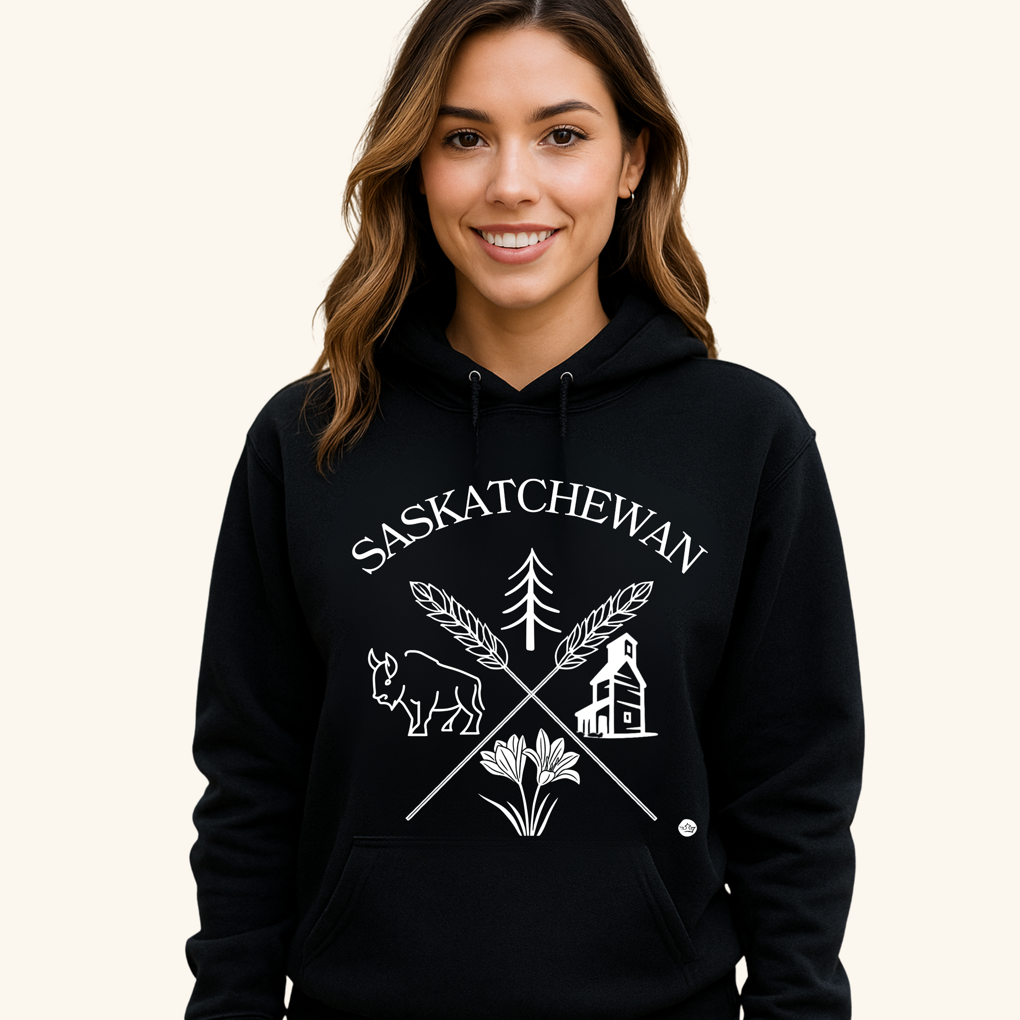 A young man with short dark hair wears a black hoodie featuring a white “Saskatchewan Prairie Roots” design with symbols of prairie life, including a bison, tree, wheat, and grain elevator. He stands with a neutral expression against a soft cream-colored background.