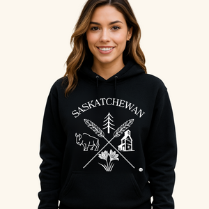 A young man with short dark hair wears a black hoodie featuring a white “Saskatchewan Prairie Roots” design with symbols of prairie life, including a bison, tree, wheat, and grain elevator. He stands with a neutral expression against a soft cream-colored background.