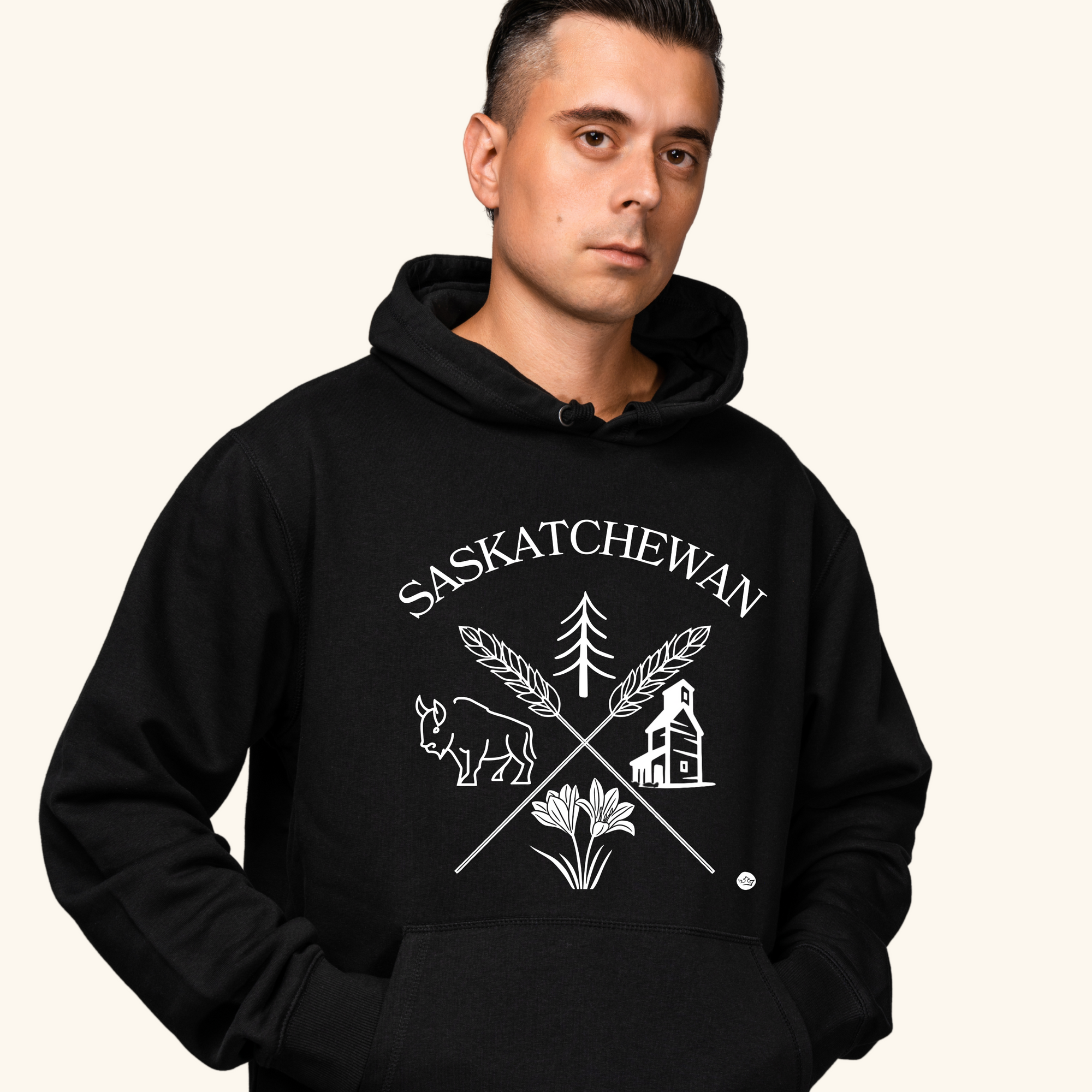 A young man with short dark hair wears a black hoodie featuring a white “Saskatchewan Prairie Roots” design with symbols of prairie life, including a bison, tree, wheat, and grain elevator. He stands with a neutral expression against a soft cream-colored background.