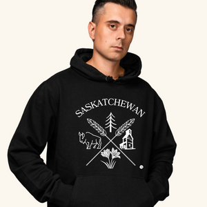A young man with short dark hair wears a black hoodie featuring a white “Saskatchewan Prairie Roots” design with symbols of prairie life, including a bison, tree, wheat, and grain elevator. He stands with a neutral expression against a soft cream-colored background.