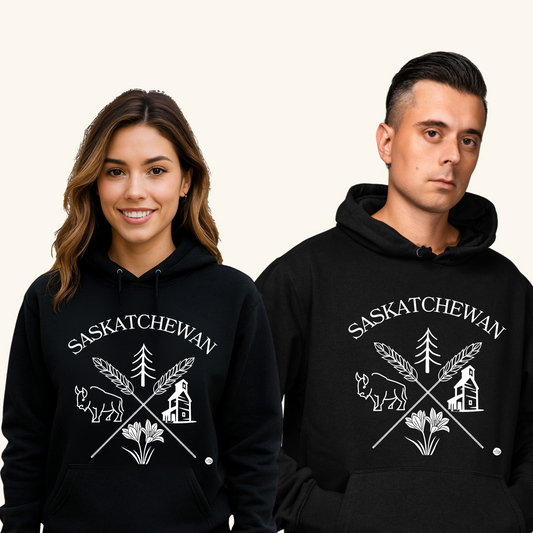 A smiling young woman and a serious-looking man stand side by side wearing matching black “Saskatchewan Prairie Roots” hoodies. The hoodie features white prairie-themed graphics including a bison, wheat stalks, prairie lily, evergreen tree, and grain elevator. They pose against a light beige background.
