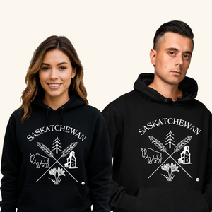 A smiling young woman and a serious-looking man stand side by side wearing matching black “Saskatchewan Prairie Roots” hoodies. The hoodie features white prairie-themed graphics including a bison, wheat stalks, prairie lily, evergreen tree, and grain elevator. They pose against a light beige background.
