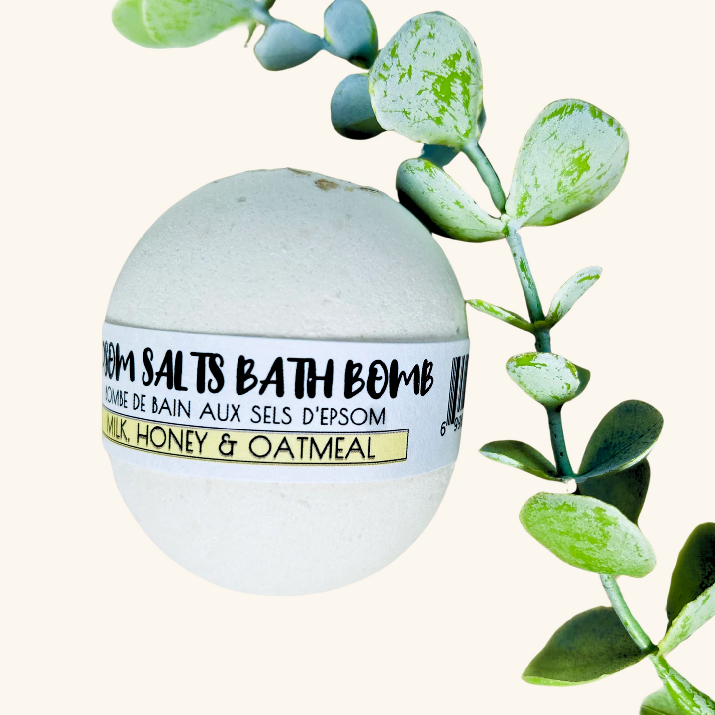 White Epsom salts bath bomb labeled Milk, Honey & Oatmeal by The Sask Bath Co., nestled among green eucalyptus leaves.