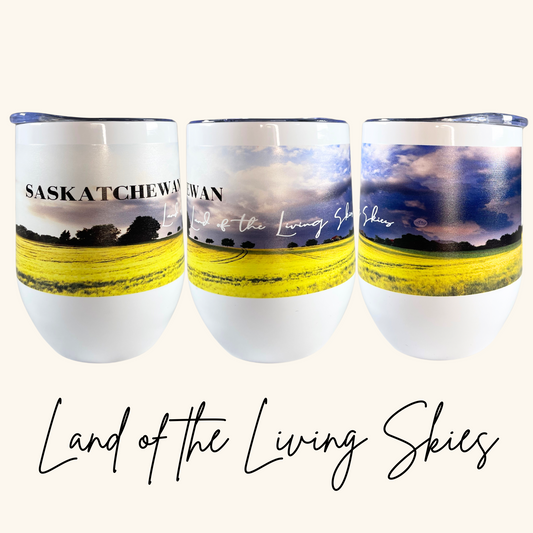 A double-walled stainless steel wine tumbler with a Prairie-inspired design, featuring intricate artwork representing the landscapes of Saskatchewan and Manitoba. The tumbler is placed on a wooden deck with a glass of wine nearby.