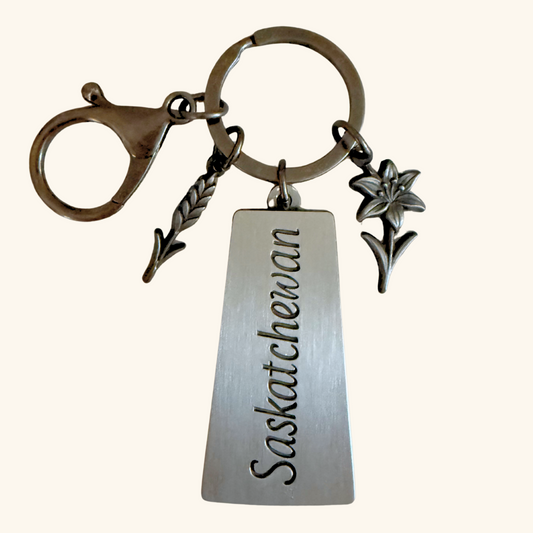 Metal keychain with a Saskatchewan-shaped engraved tag, wheat stalk charm, and Prairie Lily charm on a keyring with lobster clasp.