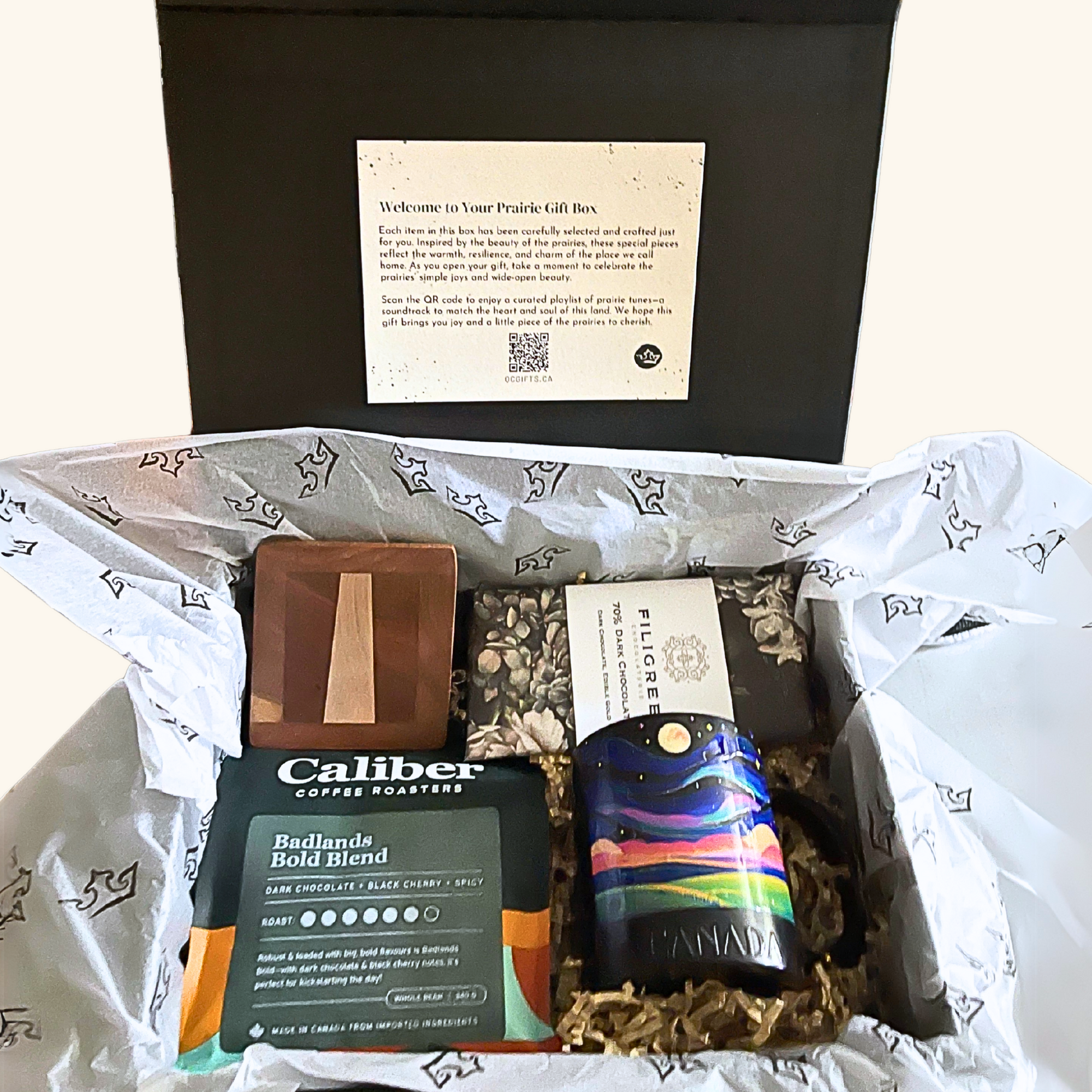 Full view of the Sunrise on the Prairie Box, showing all items inside a black magnetic gift box with a welcome card attached to the lid. Contents include Caliber Coffee, Filigree chocolate bar, a handmade wooden coaster, and a Northern Lights mug. Tissue paper with prairie motifs lines the box.