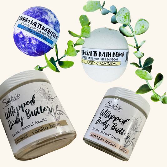 Flat lay of The Sask Bath Co. products including Coconut Vanilla Bean and Okanagan Peach Bellini body butters, and two Epsom salts bath bombs in Saskatoon Berry and Milk, Honey & Oatmeal scents, accented with eucalyptus sprigs.