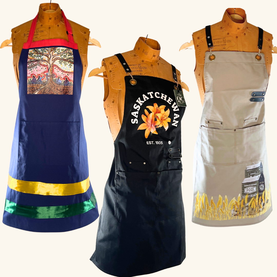 Collection of three handcrafted Prairie-inspired aprons, featuring the Saskatchewan & Prairie Lily design, Wheat Field & Grain Elevator canvas apron, and Leah Dorion’s 'Singing to the Aspen' apron