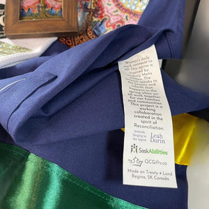 Close-up of the Singing to the Aspen apron label, highlighting the SaskAbilities partnership and hand-sewn ribbon details.